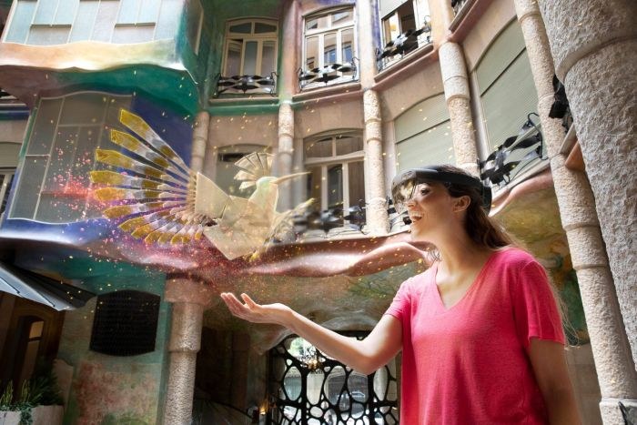 A visitor wearing HoloLens 2 in the Patio de las Mariposas holds out her hand to the paper hoopoe hologram, among Gaudí's façades